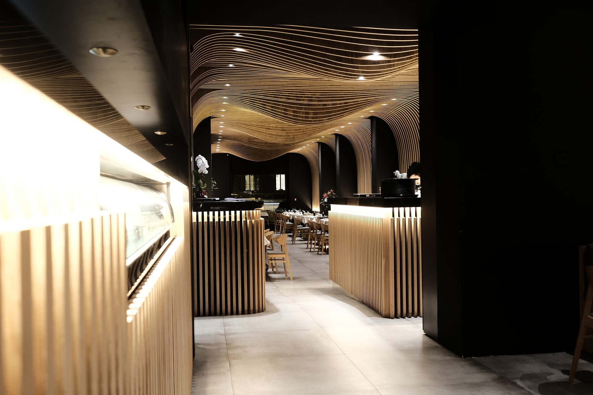 Aji Tei — corridoio interno, apertura su sala con onde di legno e cucina a vista
